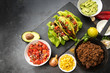 mexican tacos and ingredients like fried ground beef, tomato salsa, guacamole, corn and spices on a dark slate plate with copy space, top view from above, selected focus