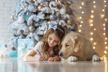 Little Cute Girl With A Golden Retriever Dog Writes Letter To Santa Near Christmas Tree