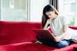 © jumlongch - Asian teenage girl using a laptop computer to check her orders online and drinking coffee on a red sofa in her home. She stressed that the sales slump.
