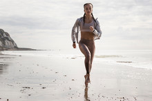 Front View Of Young Female Runner Running Barefoot Along Beach
