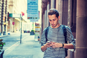 Russian Man traveling in New York, wearing striped long sleeve T shirt, shoulder carrying back bag, standing by wall on vintage street, reading, texting on cell phone, listening music with earphone..