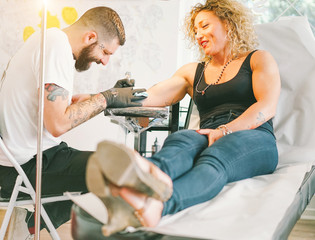 Wall Mural - Bearded tattoo artist making tattoo inside ink studio on blond woman - Tattooer at work - Contemporary trend fashion concept - Main focus on female face - Warm filter