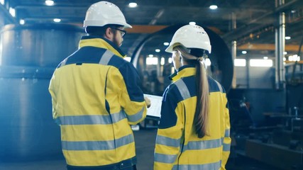 Wall Mural - Male and Female Industrial Engineers use Laptop and Have Discussion While Walking Through Heavy Industry Manufacturing Factory. They Wear Hard Hats and Safety Jackets. 