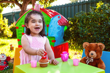Happy Laughing Baby Toddler Girl In Outdoor Second Birthday Party Holding Candle. Best Friend Teddy Bear, Playhouse And Tea Set. Pink Dress And Crown