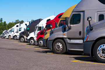 Big rigs semi trucks of different makes and models stand in row on truck stop parking lot