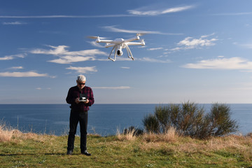 Standing on a cliff above the sea man over 50 years of age with remote control in his hands runs a white drone in the sky