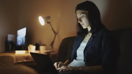 Young concentrated woman working at night using laptop computer and typing message