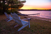 Summer Vacation At The Lake. Sunset Over A Lake With A Row Of Adirondack Chairs, Dock And Aluminum Rowboat.