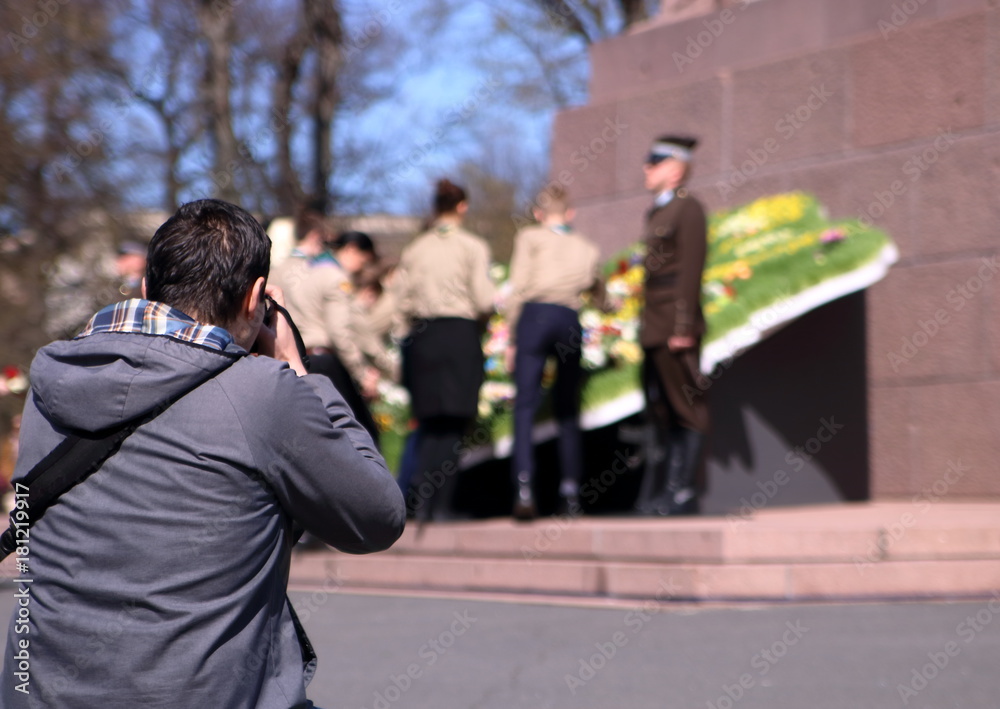 Obraz na płótnie Fotograf, tyłem, fotografuje miejsce pamięci w Rydze, Łotwa, podczas uroczystości święta narodowego, w tle niewyraźny żołnierz pełniący wartę przy mapie Łotwy zrobionej z kwiatów, młodzież w salonie