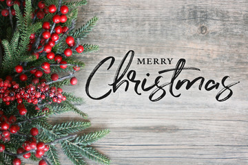 merry christmas text with christmas evergreen branches and berries in corner over rustic wooden back