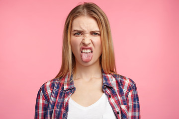 Dissatisfied female model frowns face, has disgusting expression, shows tongue, expresses non compliance, irritated with somebody, rejects do something. People and negative facial expressions