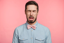 Bearded Brunet Attractive Man Closes Eyes And Opens Mouth Sees Something Sour, Isolated Over Pink Background. Unhappy Handsome Young Male Wears Pink Bow Tie And Shirt, Makes Grimace Or Pulls Face