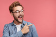 Excited surprised fashionable guy with trendy hairstyle and beard points happily aside on copy space of pink background for your fashion clothing advertising information or promotional text.