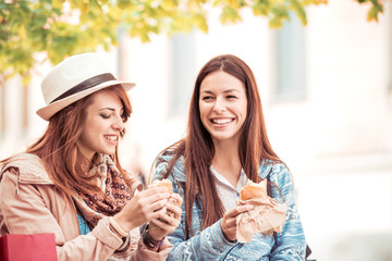 Two young female friends laugh and eat sandwich outdoors.