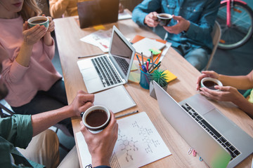 Poster - Top view close up group of colleagues holding mug of appetizing beverage while working at desk in modern office. Occupation concept