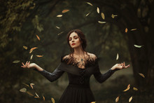 Incredible, Amazing, Seductive Girl, In A Black Dress , Magic Rotates The Leaves. Background Is Fantastic Autumn. Artistic Photography. Fantasy Gothic Woman Dark Queen. Lady Medieval Witch