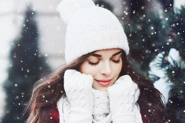 Outdoor close up portrait of young beautiful happy smiling girl wearing white knitted beanie hat, scarf and gloves. Model posing in street. Winter holidays concept. Toned