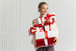 © deniskomarov - Blond hair woman in white sweater holding a lot of presents over white wall background indoor. Smiling woman holding paper gift boxes red color and looks at camera. Toothy smiling woman's face.