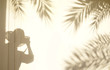 Conception for exotic travel. The shadow of the leaves of a palm tree and a photographer in a hat on a light wall at sunset or sunrise. Template background for text, wallpaper.