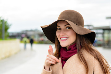 Wall Mural - Attractive woman in a brown hat and beige coat looking at the camera points the finger at you and smiles. Pick me.