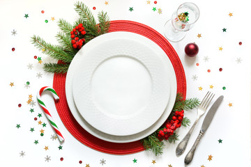 Christmas table setting with white dishware, silverware and red decorations on white background. Top view.