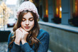 Outdoor close up portrait of young beautiful girl with long hair wearing pink big loop hat, blue sweater posing in street of european city.  Christmas, winter holidays concept. Copy, empty space 