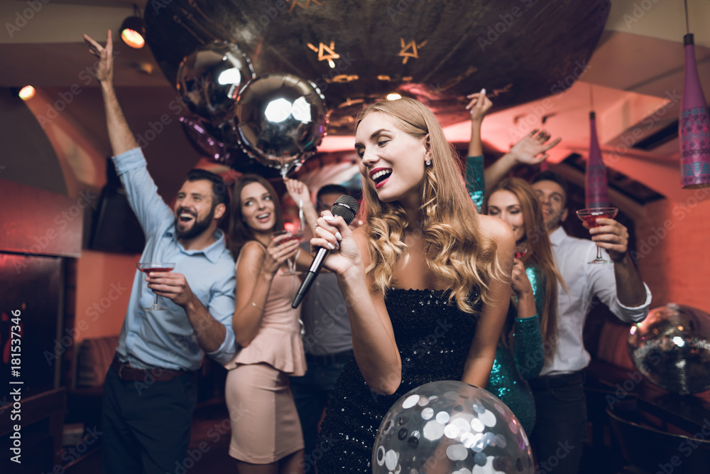 Young people have fun in a nightclub and sing in karaoke. In the foreground there is a woman in a black dress. - obrazy, fototapety, plakaty 