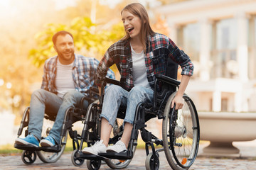 A man and a woman on wheelchairs ride around the park. They arranged a race in wheelchairs.