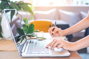 Woman hand works in a laptop computer and writing on notepad with a pen in the office.