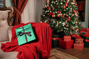 Green christmas gift close-up on foreground. Bow of red ribbon. Abstract background with blurred lights and tree. copy space