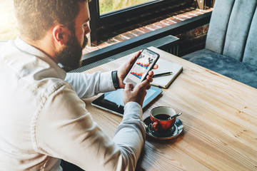 Wall Mural - Back view.Young bearded businessman in white shirt is sitting at table,using smartphone with graphs,charts,diagrams on screen.On table is tablet computer,notebook.Online marketing,education,e-learning