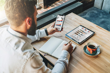 Wall Mural - Back view.Young bearded businessman in white shirt is sitting at table,using smartphone with graphs, charts and diagrams on screen and making notes in notebook.Online marketing,education, e-learning.