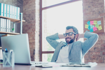 Portrait of young, smiling, attractive economist in glasses and jacket, sitting at his desktop holding hand behind the head, watching something on computer, having recreation from work