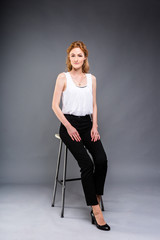 beautiful young caucasian woman with long red hair in high heels sitting on a chair, black trousers and white shirt in full growth on a gray background. Business concept