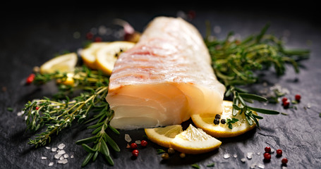 Fresh raw cod fillet with addition of herbs and lemon slices on black stone background