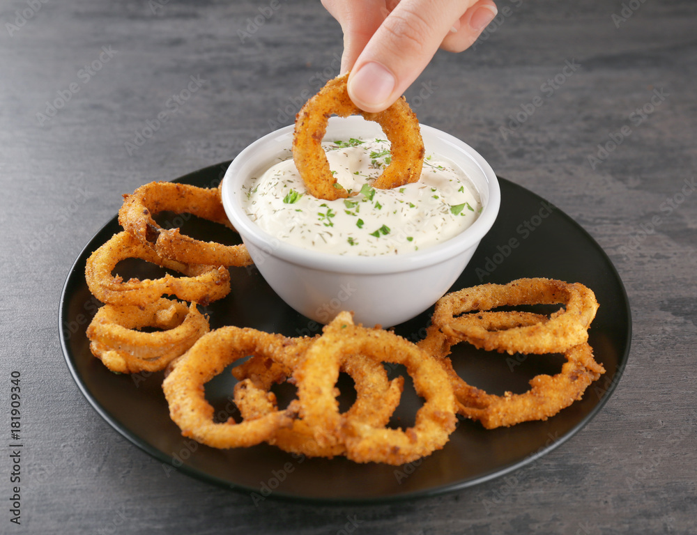 Woman dipping onion ring into bowl with sauce on table