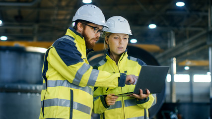 Wall Mural - Male and Female Industrial Engineers in Hard Hats Discuss New Project while Using Laptop. They Make Showing Gestures.They Work at the Heavy Industry Manufacturing Factory.