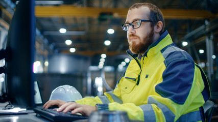 Wall Mural - Industrial Engineer Works at Workspace on a Personal Computer.  He Wears  Safety Jacket and Works in the Main Workshop of the Heavy Industry Manufacturing Factory.