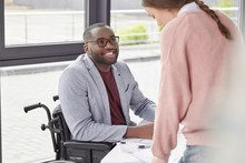 Disable Dark Skinned Male Sits On Wheelchair, Looks Happily At His Secretary Who Presents Information, Discuss Opinions And Ideas, Give Advice For Each Other, Speak About Details Of Working Process