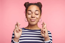 Isolated Shot Of Attractive Coquette Female Model With Dark Skin Closes Eyes And Crosses Fingers In Anticipation, Believes For Good Luck, Isolated Over Pink Studio Background. Woman Gestures Indoors