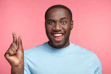 Cheerful dark skinned male with happy expression shows something tiny, opens mouth widely, shows white perfect teeth, isolated over pink background. African positive guy gestures in pink studio