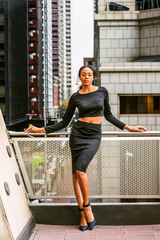 African American Business Woman Fashion in New York. Lady wearing black long sleeve crop top, skit, high heel pumps shoes, stands by railing on balcony. Street with high buildings on background..