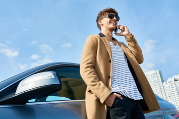 Wall Mural - Low angle portrait of handsome young man speaking by phone standing in sunlight leaning on car outdoors against clear blue sky, copy space