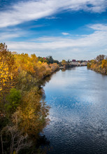 River View With Trees In Autumn Colors On Both Banks. A Bridge Is Crossing The River In The Distance. A Blue Sky With Clouds Is In The Background. The Water Is Reflecting The Blue Sky And White Clouds