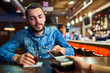Portrait of drunk young man paying via smartphone buying drinks in bar, focus on payment terminal in foreground
