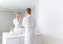 Back View Of Confident Girl Applying Cream On Her Cheek While Standing In Front Of The Mirror At Spa