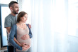 © Yakobchuk Olena - Soon we will become parents. Excited young family is looking over the window with joy and smiling. Man is embracing pregnant woman with affection. Copy space