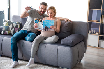 Wall Mural - Planning future journey. Full length portrait of cheerful loving couple reading magazine about traveling while sitting on sofa at home. Man is holding plane toy while his pregnant wife is smiling