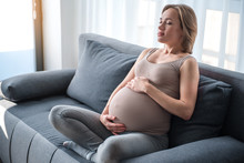 Stay Calm And Relax. Portrait Of Serene Pregnant Woman Meditating At Home. She Is Sitting On Couch And Touching Her Belly With Love. Her Eyes Are Closed With Tranquility