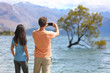 © Maridav - New Zealand tourists taking phone pictures of Wanaka Lone Tree at lake. People looking at view of famous touristic attraction in south island, Otago Region, NZ.